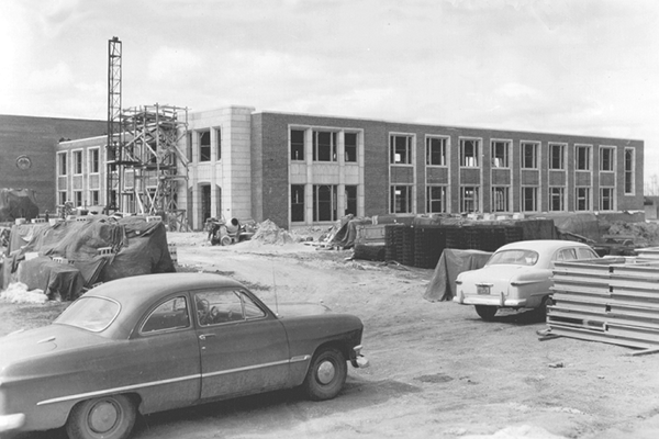Black and white photo of a partially constructed building. Building materials and tools are scattered throughout the photo and two cars from the 1940's are in the foreground.