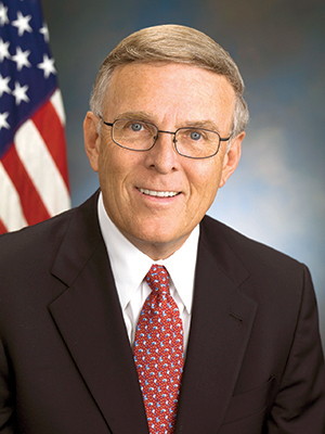 Byron Dorgan wearing a black suit and red tie with U.S. flag in background