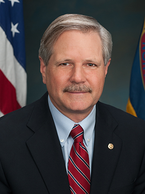 John Hoeven wearing a black suit, red tie, and gold pin